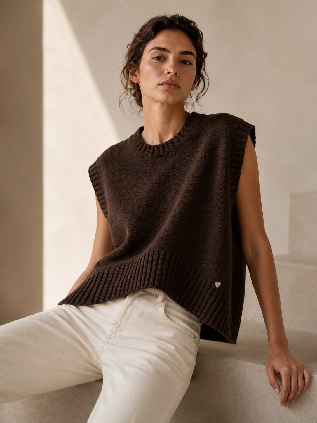 A woman wearing a sleeveless dark brown ribbed knit sweater vest and off-white trousers, seated on a light-colored stone step in a warm, minimalist interior.