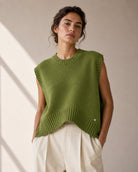 A woman wearing a sleeveless olive green knit sweater vest with ribbed trim over cream-colored trousers, standing against a neutral beige wall with soft diagonal shadows.