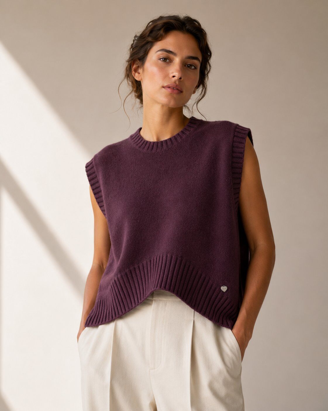 A woman wearing a sleeveless ribbed knit sweater vest in a deep plum purple, paired with off-white trousers, standing against a neutral beige wall 