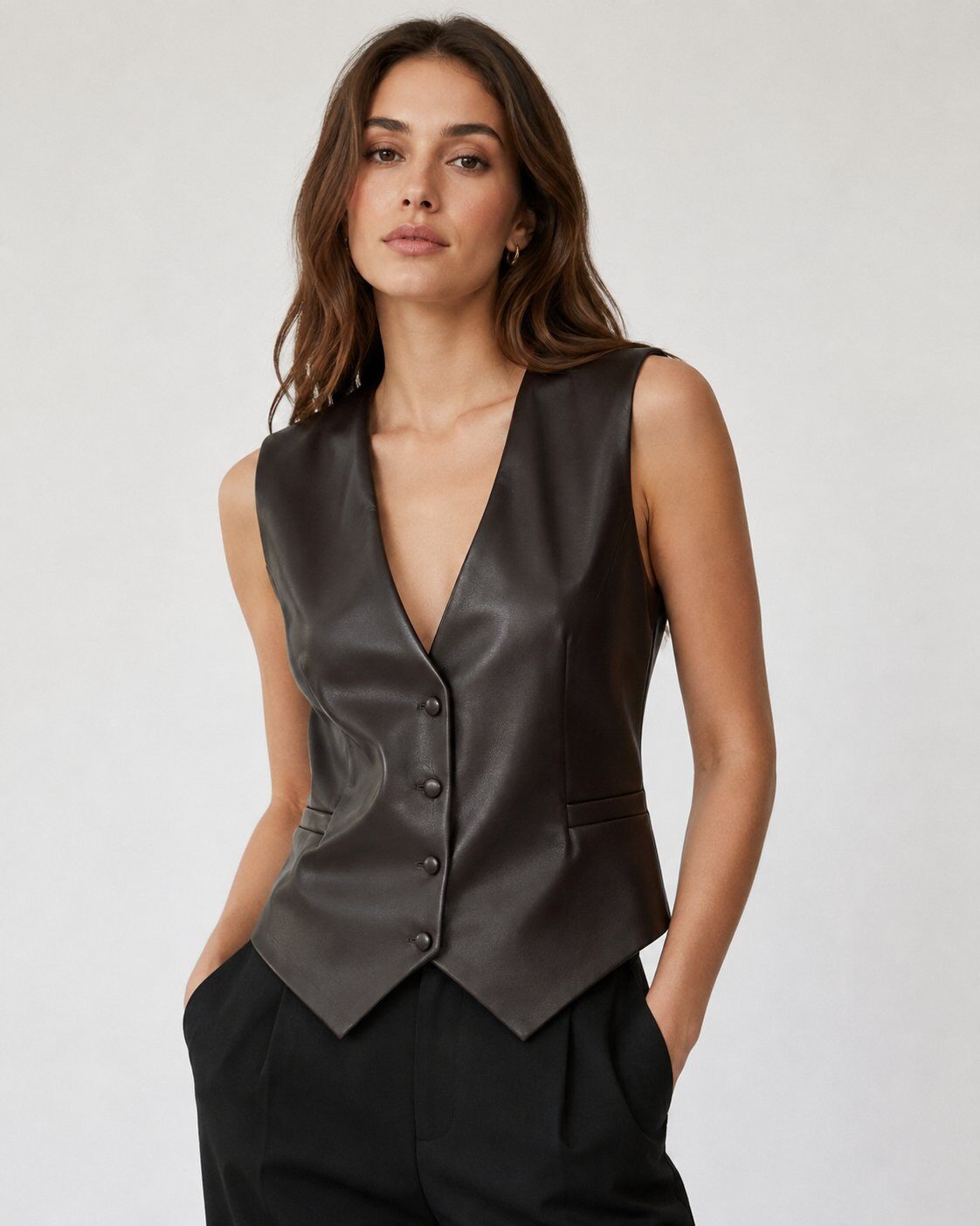 A woman modeling a dark brown leather vest with a deep V-neck and button-front closure, paired with black trousers, against a plain light gray studio background.