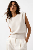 A woman wearing a sleeveless cream knit sweater vest with a ribbed hem, paired with matching cream trousers, against a clean white studio background.
