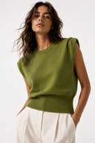 A woman wearing a sleeveless olive green knit sweater with a crew neck and ribbed hem, paired with white trousers, photographed against a clean white studio background.