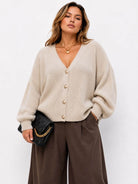 A woman wearing a beige ribbed knit cardigan with gold buttons, paired with brown wide-leg trousers and holding a black quilted clutch bag with a gold chain strap, set against a plain white background.