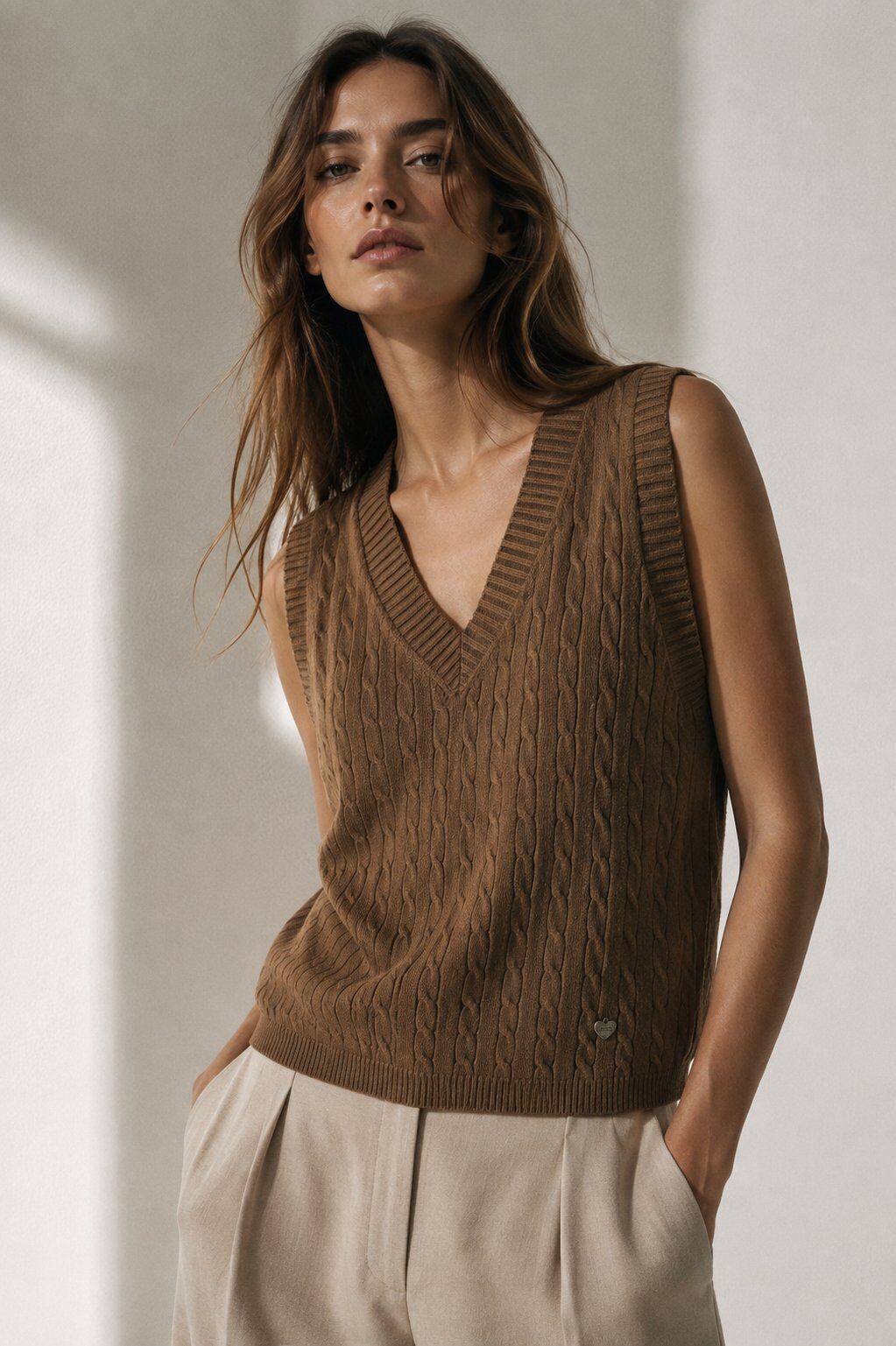 A woman wearing a brown cable-knit sleeveless V-neck sweater vest and light beige trousers, standing against a neutral light gray wall with soft shadows.