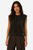 A woman modeling a sleeveless, dark brown fuzzy knit vest with a small gold button detail on the lower right, paired with dark brown wide-leg trousers, against a plain white background.