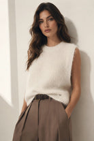 A woman wearing a cream-colored, sleeveless, ribbed knit sweater vest tucked into high-waisted taupe trousers, leaning against a white wall.