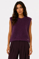 A woman modeling a sleeveless, fuzzy knit top in a deep plum purple color, paired with dark pleated trousers, against a plain white studio background.