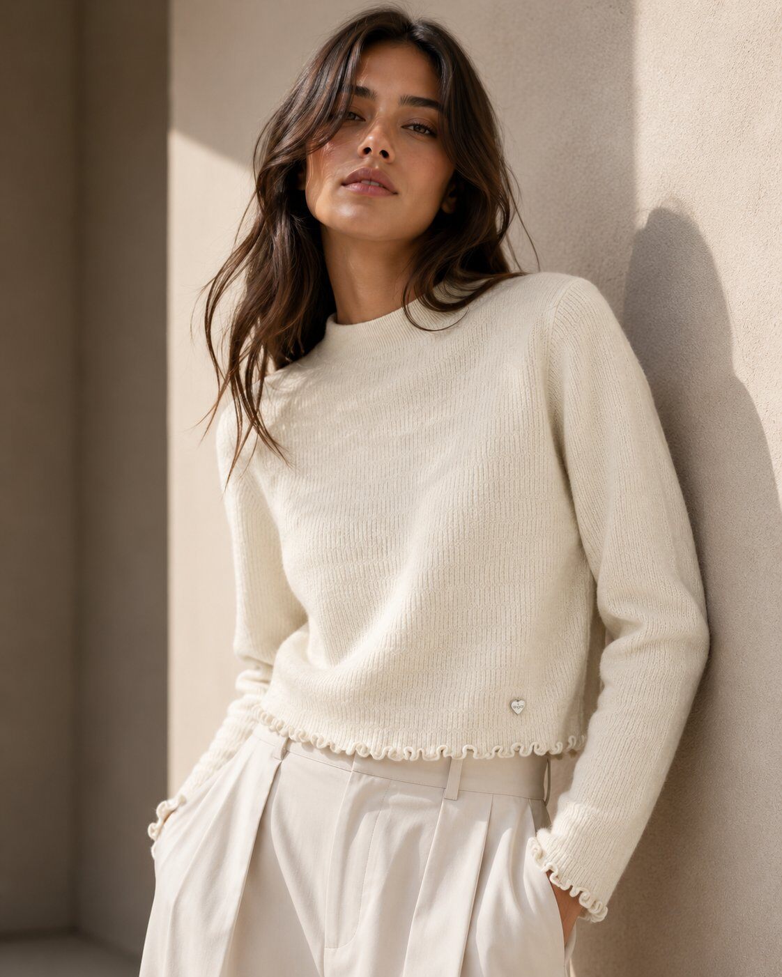 A woman wearing a cream ribbed knit sweater with delicate ruffle trim at the hem and cuffs, paired with matching cream trousers, leaning against a textured beige wall.