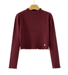 A long-sleeve ribbed knit mock-neck sweater in deep burgundy, hanging on a wooden hanger against a plain white background. The bottom hem has a scalloped edge, and a small white heart detail is visible on the lower right side.