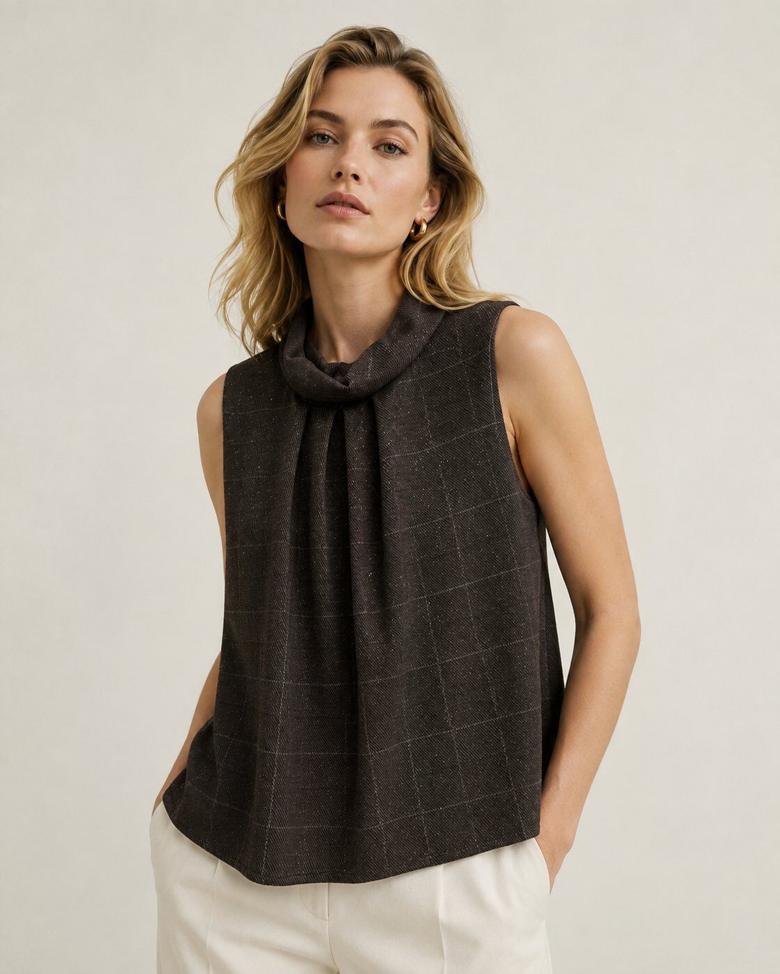 A woman wearing a dark brown sleeveless top with a cowl neck and a subtle windowpane plaid pattern, paired with off-white trousers, against a plain light beige background.