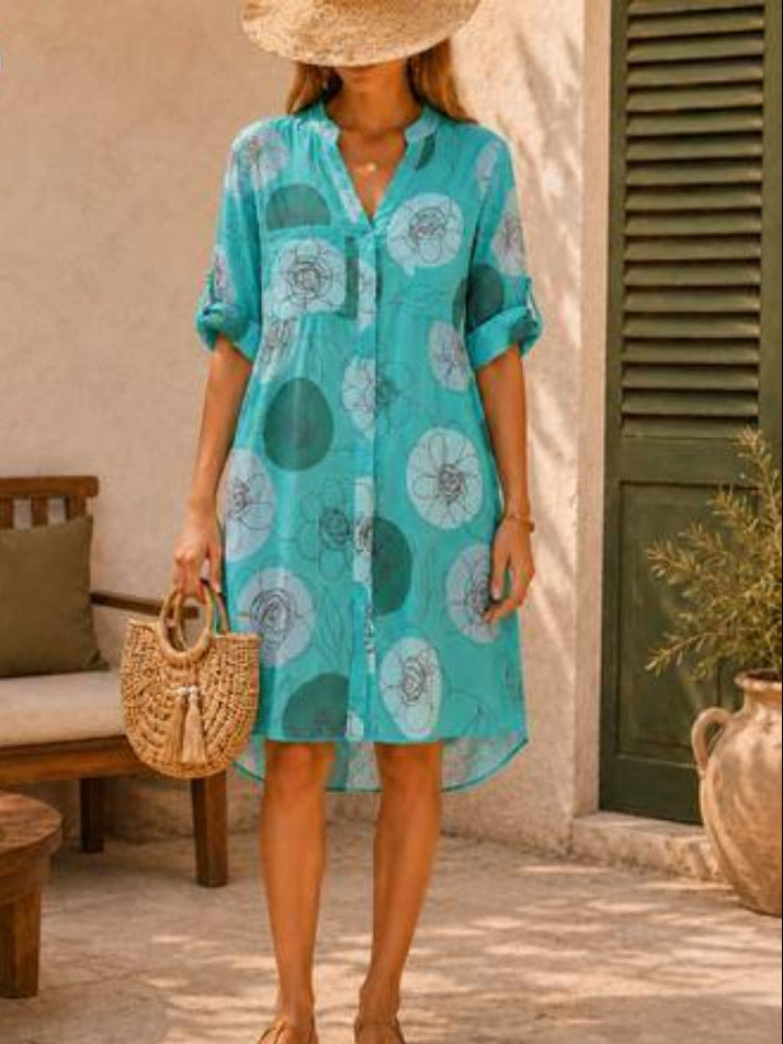 A woman wearing a turquoise blue button-up shirt dress with a white and dark green floral pattern, styled with a straw hat, a woven straw handbag, and tan sandals in an outdoor patio setting.