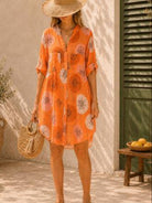 A woman wearing a short-sleeve orange button-up shirt dress with a large floral print, styled with a straw hat, woven bag, and sandals in an outdoor patio setting.