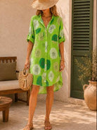 A woman wearing a bright green short-sleeve button-up dress standing in a sunlit outdoor patio setting with a woven straw hat, straw bag, and sandals.