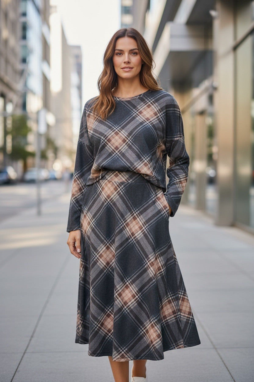 A woman stands on a city sidewalk wearing a long-sleeve, knee-length plaid dress in dark grry, beige, and brown tones.