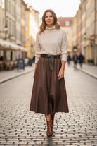 A woman stands on a cobblestone street wearing a cream turtleneck sweater, a brown midi skirt with a black belt, and brown ankle boots.