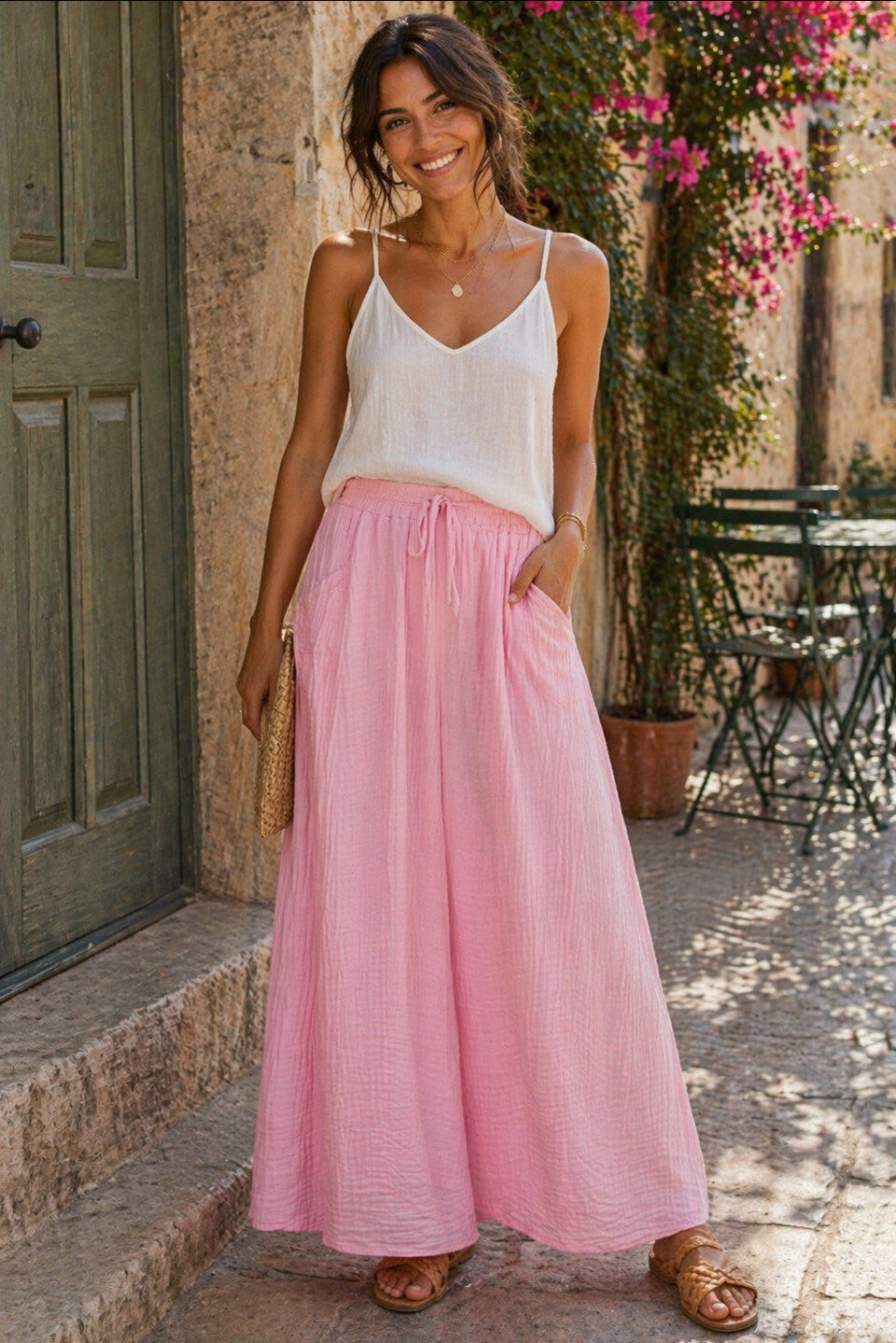 Woman wearing a white top and pink skirt standing outdoors near a building with plants.