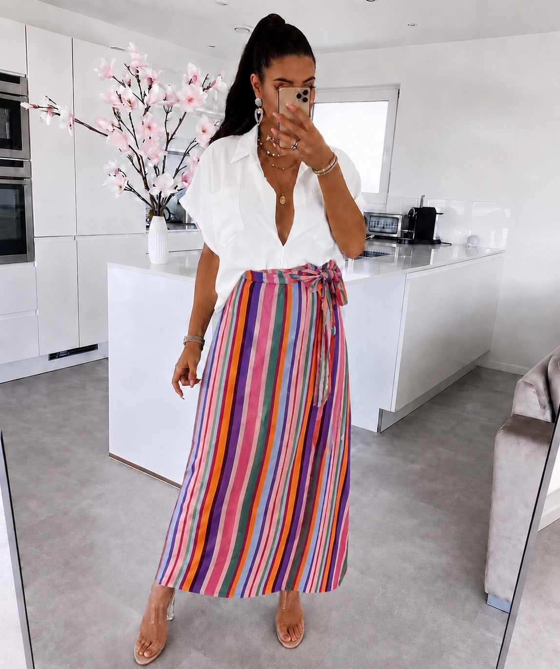 A woman posing for a mirror selfie in a modern white kitchen, wearing a white button-down shirt and a high-waisted, multicolor vertically striped wrap skirt with a tie waist.