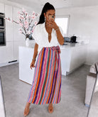 A woman posing for a mirror selfie in a modern white kitchen, wearing a white button-down shirt and a high-waisted, multicolor vertically striped wrap skirt with a tie waist.