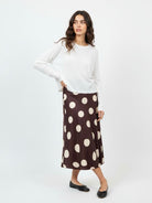 A woman modeling a white long-sleeve knit top and a brown midi skirt with large cream polka dots, paired with black flats, against a plain white studio background.