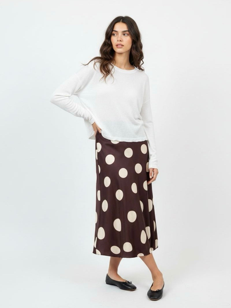 A woman modeling a white long-sleeve knit top and a brown midi skirt with large cream polka dots, paired with black flats, against a plain white studio background.