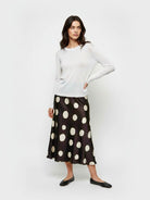 A woman modeling a white long-sleeve knit top and a black midi skirt with large cream polka dots, paired with black flats, against a plain light gray studio background.