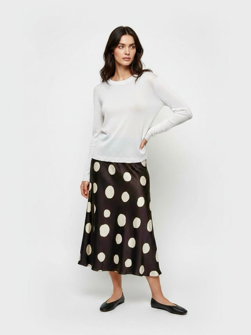 A woman modeling a white long-sleeve knit top and a black midi skirt with large cream polka dots, paired with black flats, against a plain light gray studio background.