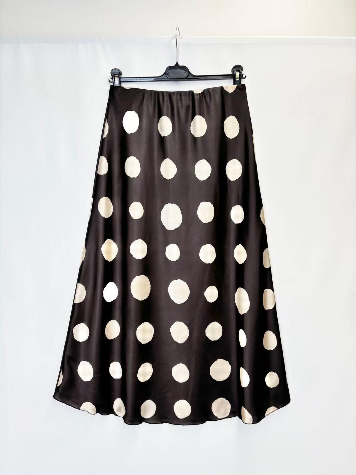 A black satin midi skirt with large cream polka dots, hanging on a black hanger against a white background.