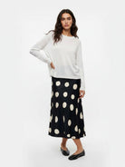 A woman modeling a white long-sleeve knit top and a black midi skirt with large cream polka dots, paired with black flats, against a plain white studio background.