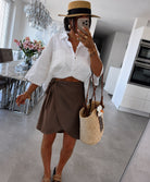 A woman wearing a white cropped button-up shirt, a brown wrap-style mini skirt