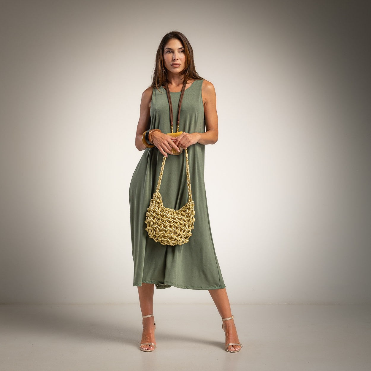 A woman modeling a sleeveless olive green midi dress, holding a chunky woven tan shoulder bag