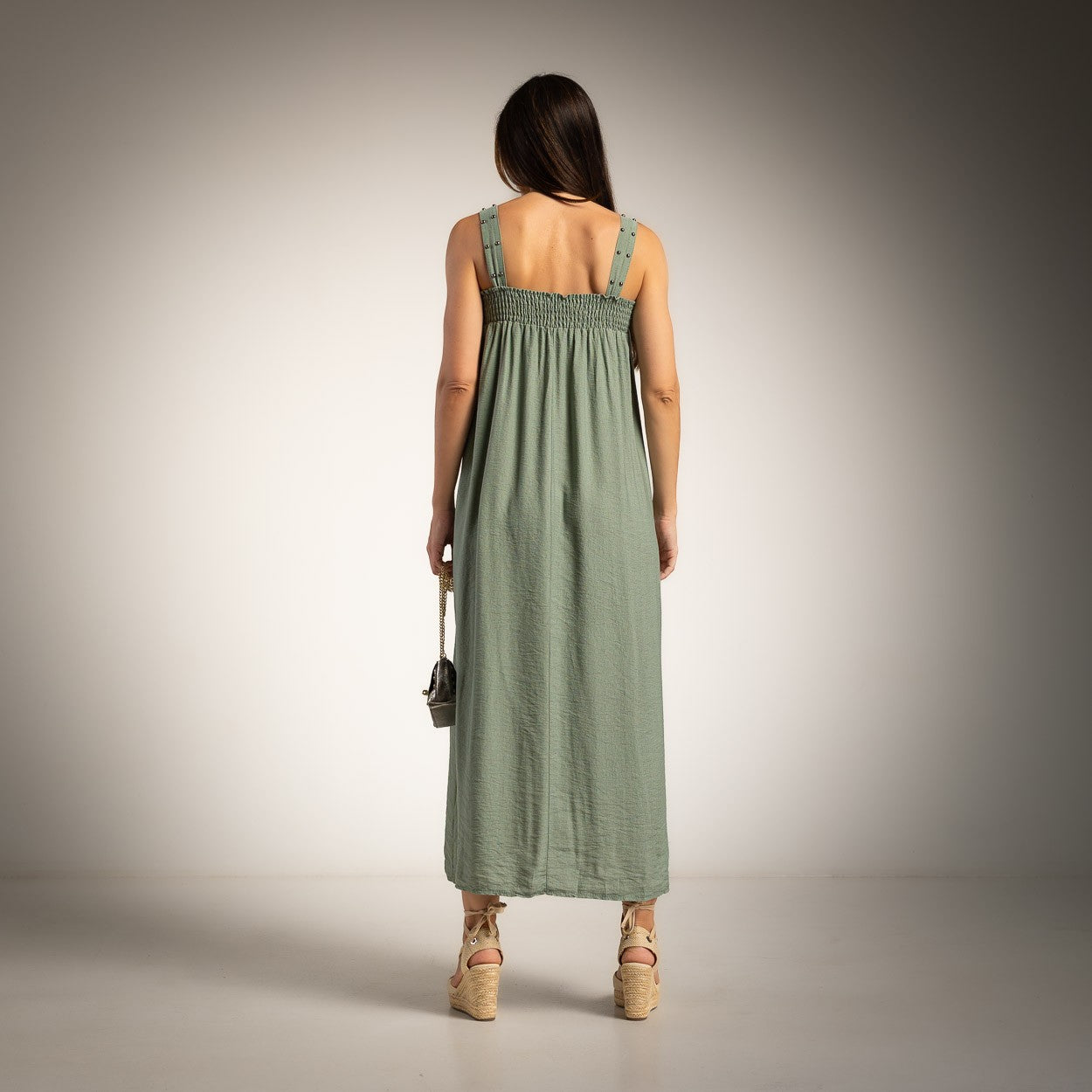 A woman viewed from behind wearing a green sleeveless maxi dress with a smocked bodice and straps