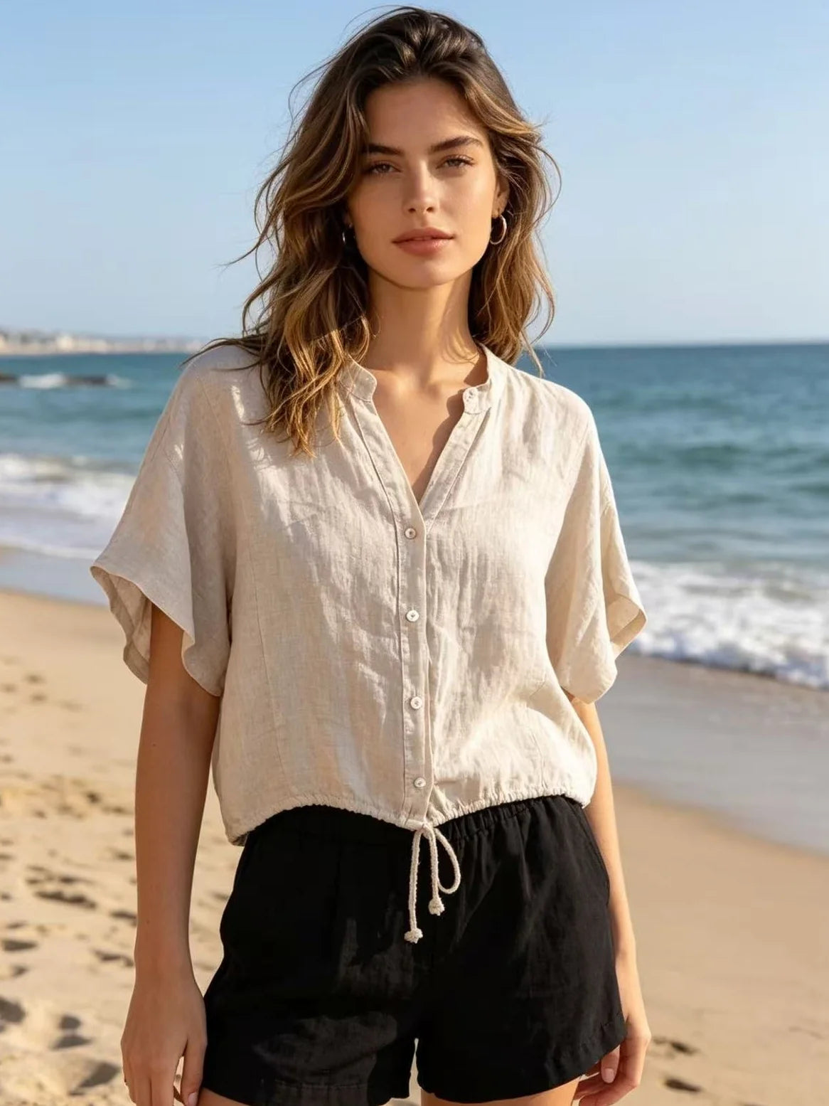 A woman standing on a sandy beach wearing a light beige short-sleeve linen button-up shirt and black drawstring shorts.