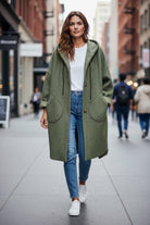 A woman walking down a city street wearing a long khaki green textured coat over a white t-shirt, blue jeans, and white sneakers.