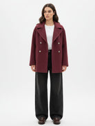 A woman stands wearing a burgundy double-breasted coat over a white top and black wide-leg trousers.