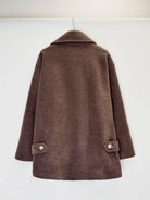 A brown wool-blend coat with a wide collar and buttoned flap pockets on the sleeves, hanging against a plain white wall.