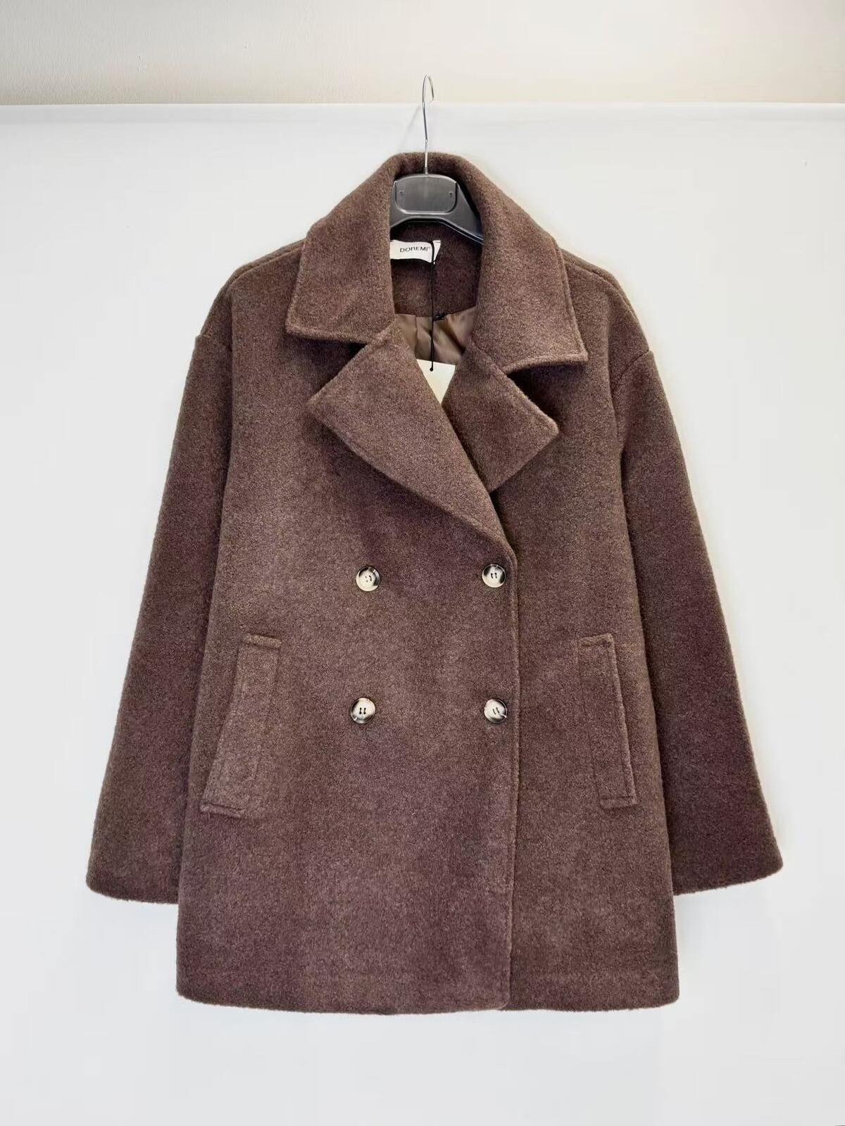 A brown double-breasted coat with wide lapels and buttons, hanging on a black hanger against a plain white wall.