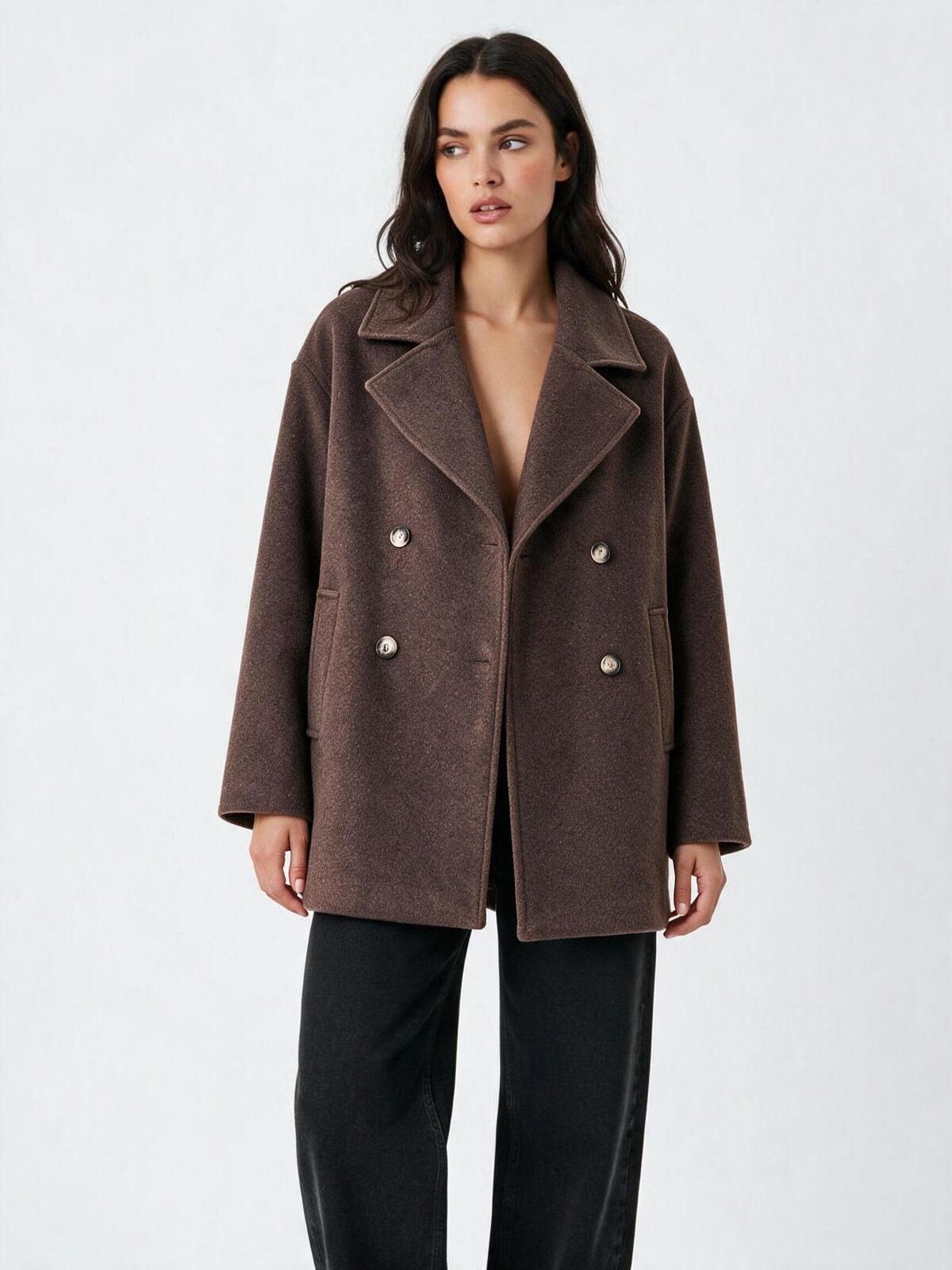 A woman modeling a brown double-breasted coat with silver buttons, paired with black trousers, against a plain light gray studio background.