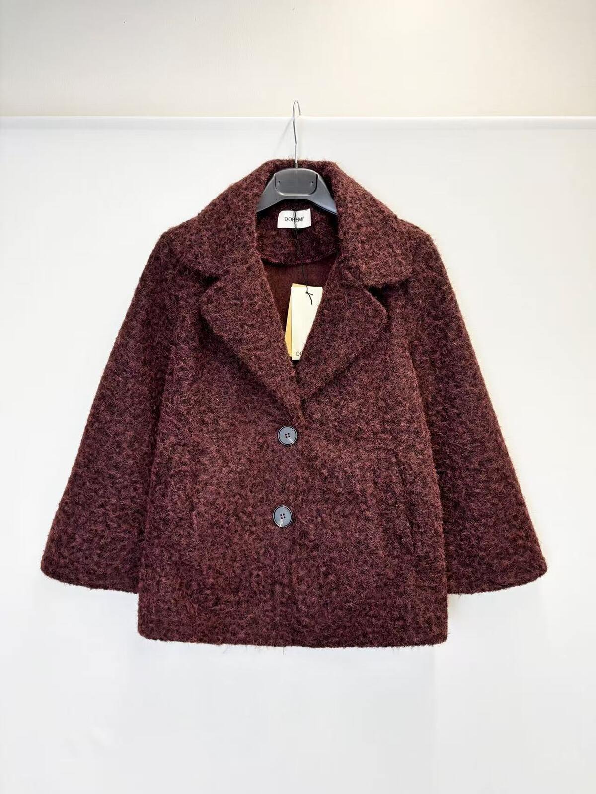A textured, boucle-style dark burgundy coat with wide lapels and two front buttons, hanging on a black hanger against a plain white wall.