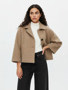 A woman modeling a cropped, textured beige coat with wide three-quarter sleeves, worn over a white turtleneck and black jeans, against a plain white studio background.