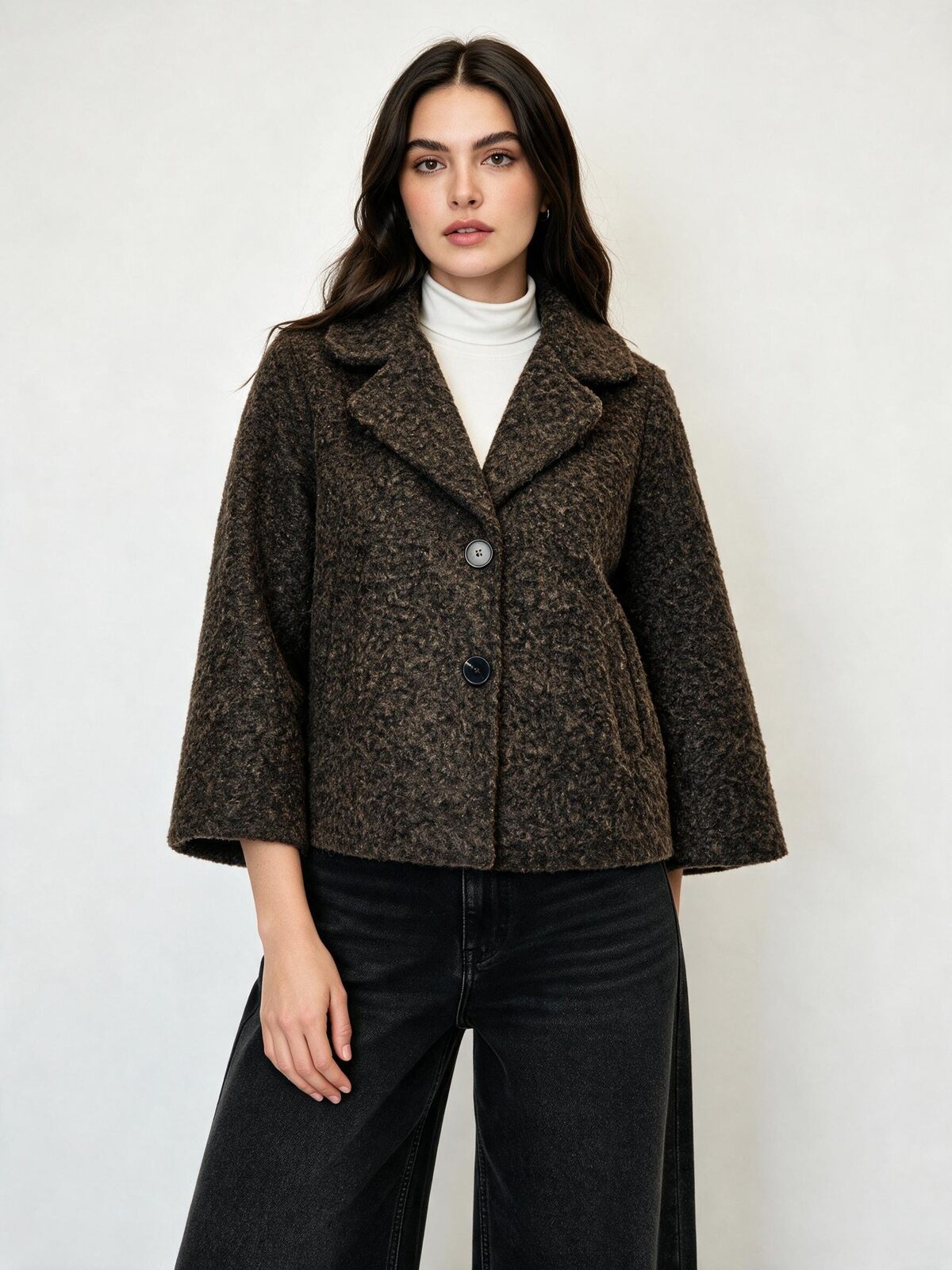 A woman modeling a cropped, dark brown textured tweed jacket with wide three-quarter sleeves, worn over a white turtleneck and black wide-leg jeans, against a plain light gray background.