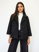 A woman modeling a dark charcoal gray textured wool-blend coat with a white turtleneck and black wide-leg jeans against a plain white studio background.