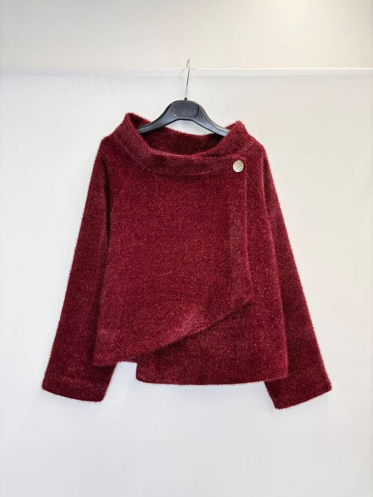 A textured, deep burgundy long-sleeve sweater with an asymmetrical wrap-style hem and a single decorative button, hanging on a black hanger against a plain white wall.