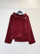 A textured, deep burgundy long-sleeve sweater with an asymmetrical wrap-style hem and a single decorative button, hanging on a black hanger against a plain white wall.