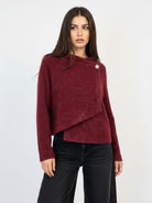 A woman modeling a textured, off-the-shoulder burgundy knit sweater with a single gold button detail, paired with black jeans, against a plain white studio background.