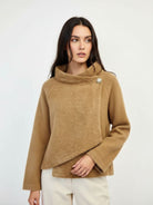 A woman wearing a camel-colored knit sweater with a cowl neck and asymmetrical button detail, paired with cream pants, against a plain light gray background.