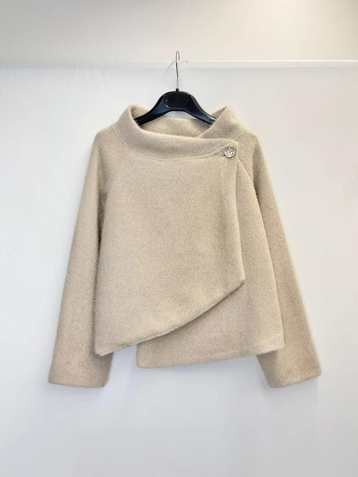 A beige textured knit sweater with a high mock neck and asymmetrical draped front detail, hanging on a black hanger against a plain white wall.
