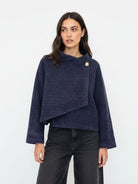 A woman modeling a navy blue textured knit wrap jacket with a single gold button closure, paired with black jeans, against a plain white studio background.