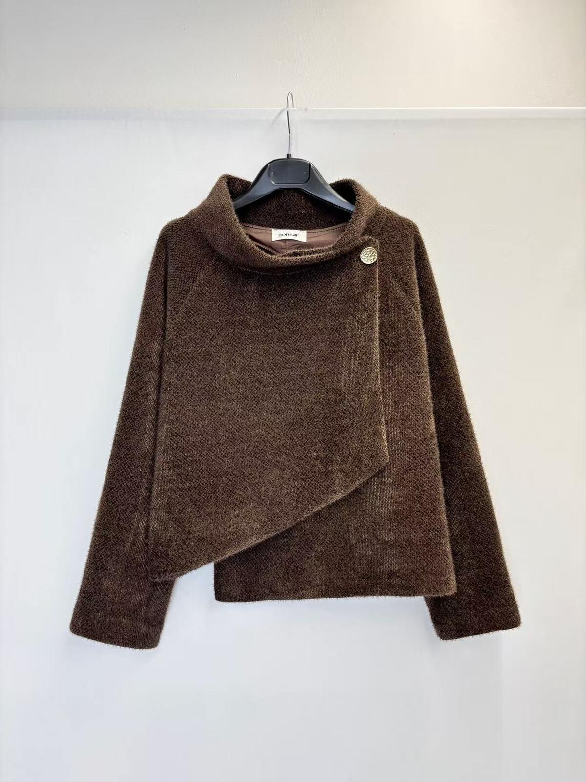 A brown textured wrap jacket with a high mock neck and asymmetrical button detail, hanging on a black hanger against a plain white wall.