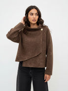 A woman modeling a brown textured knit asymmetrical wrap jacket with a single decorative gold button, paired with black wide-leg jeans, against a plain white studio background.