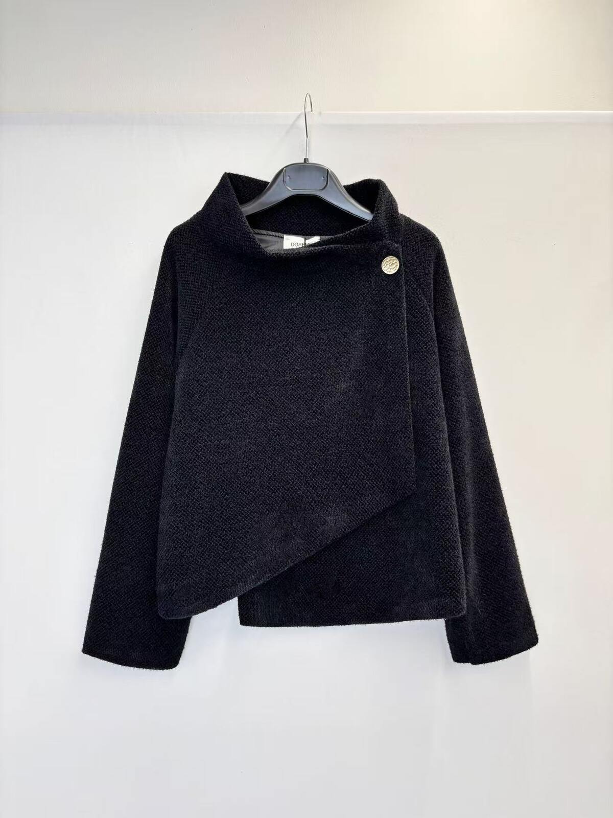A black textured wrap-style jacket with a high mock neck and asymmetrical button closure, hanging on a black hanger against a plain white wall.